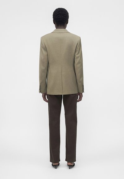 Άτομο που στέκεται πλάτη φοράει μπεζ σακάκι, σκουρό καφέ παντελόνια και μαύρα παπούτσια με τακούνι σε μονόχρωμο φόντο.