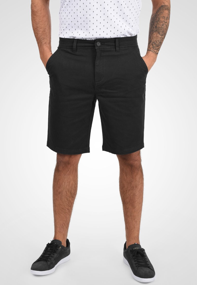 Schwarze Baumwollshorts mit klassischem Schnitt und flachem Bund. Verfügt über Seitentaschen und einen Knopfverschluss. Getragen mit schwarzen Turnschuhen.