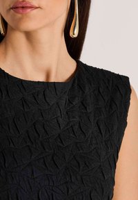 Schwarzes, strukturiertes Kleid mit geometrischem Muster, rundem Ausschnitt und ärmellosem Design. Goldene Tropfhänger schmücken das Outfit.