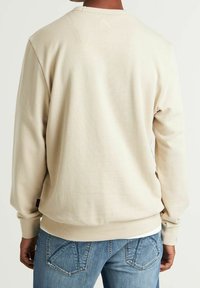 Felpa beige con texture a coste e scollatura rotonda, caratterizzata da cuciture visibili e un discreto dettaglio del logo sul retro, abbinata a jeans blu.