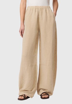 Beige bukser med vide ben og elastisk talje, båret med sorte sandaler, mod en neutral baggrund.