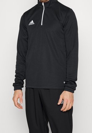 Personne portant un haut noir Adidas à manches longues avec fermeture quart-zip et un pantalon noir assorti avec le logo Adidas sur la poitrine et la cuisse.