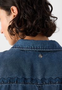 Mørkeblå denimjakke med spiss krage, sikksakk-søm langs yoken og en metallknapp bak. Teksturert stoff er synlig.