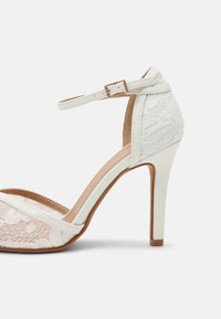 Sandale blanche à talons hauts avec surcouche en dentelle, bride à la cheville et boucle dorée, vue de profil sur fond blanc uni.