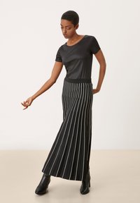 Zwarte satijnen top met korte mouwen in combinatie met een zwart-wit verticaal gestreepte, geplisseerde maxi rok. Zwarte enkellaarsjes maken de look compleet.