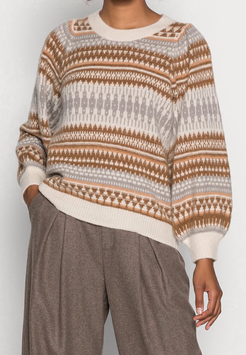 Personne portant un pull en maille marron, blanc et beige à motifs avec des manches bouffantes et un pantalon marron à jambes larges.