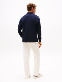 Camisa polo de manga longa azul-marinho com gola, calças brancas de corte confortável e sapatilhas brancas; cabelo cacheado visível por trás.