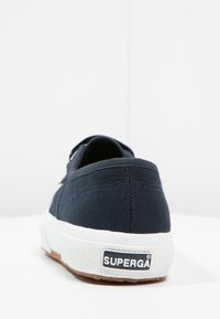 Zapatilla de lona azul oscuro con suela de goma blanca, con un acabado texturizado y un logo de Superga en el talón, que muestra un diseño minimalista.