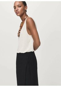 Top sin mangas beige claro combinado con una falda larga negra de envoltura que presenta un detalle de nudo, con un collar de cuentas de madera gruesas que realza el conjunto.