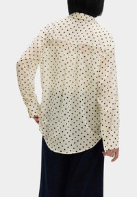 Camicia a pois color crema, con pois neri. Presenta maniche lunghe, colletto a punta e orlo arrotondato, realizzata in un tessuto leggero.