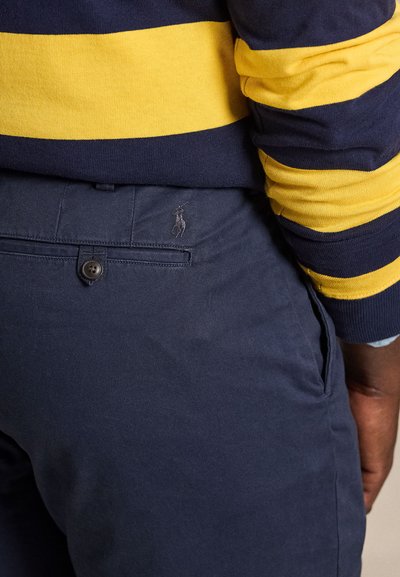 Person wearing navy blue pants with buttoned back pocket and navy and yellow striped long-sleeve shirt, arm relaxed by side.