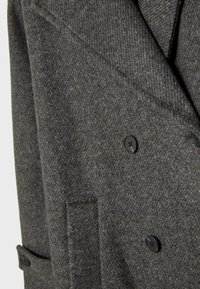 Kaksirivinen harmaa takki, joka on valmistettu teksturoidusta kankaasta ja jossa on houndstooth-kuvio. Takissa on mustat napit ja sivutasku.