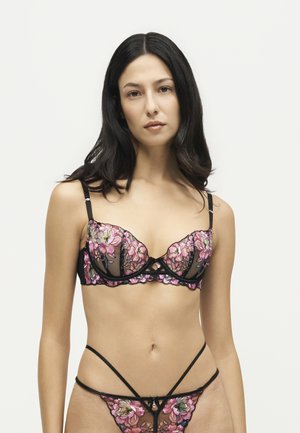 Woman with long dark hair wears black sheer lingerie with pink floral embroidery against a plain light background.