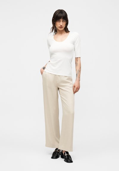 Mujer con cabello oscuro, vestida con una camiseta blanca de cuello redondo, pantalones anchos beige y mocasines negros, de pie frente a un fondo claro y sencillo.