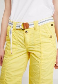 Pantalon jaune à finition texturée, doté d'une ceinture rayée blanche et grise et d'accents en cuir marron, fermé par des boutons métalliques.