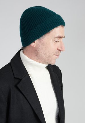 Gebreide teal beanie met een geribde textuur, gedragen met een witte coltrui en een zwarte jas; zijprofielweergave benadrukt de pasvorm en kleur.