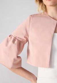 Vaaleanpunainen lyhyt takki, jossa on pomppivat hihat, sileä tekstuuri ja ilman sulkijoita. Ominaisuuksiin kuuluu leveä kaula-aukko ja minimalistinen muotoilu.