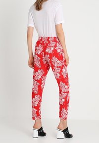 Vrouw die hoge rode broek met witte bloemenprint draagt, een witte blouse met korte mouwen en zwarte open teen blokhakken, staande met haar rug naar de kijker.