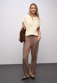 Haut en maille crème clair à manches courtes, pantalon taille haute en satin marron, et ballerines pointues noires. Sac en daim marron avec poignées latérales.