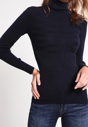 Pull à col roulé côtelé bleu marine avec des rayures horizontales discrètes porté avec un jean bleu foncé, modèle posant les mains sur les hanches.