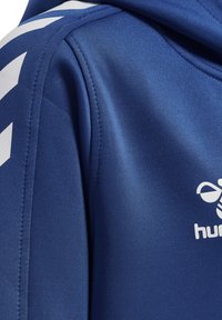 Felpa sportiva blu con tessuto morbido, logo bianco e motivo a chevron sulle maniche. Il design include un cappuccio con cordino per una vestibilità regolabile.