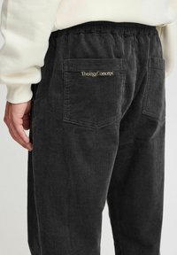 Černé kordové kalhoty s žebrovanou strukturou, se zadní kapsou zdobenou zlatou výšivkou "The/oggConcept." Viditelný elastický pas.