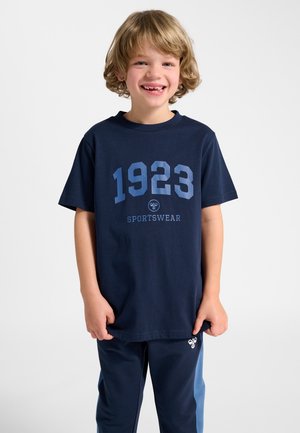 Lächelnder blonder Junge trägt ein marineblaues "1923 Sportswear" T-Shirt und passende Hose mit hellblauen Seitenstreifen vor weißem Hintergrund.