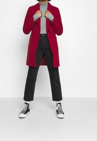 Cappotto lungo rosso in misto lana, con chiusura a un bottone e due tasche a toppa. Abbinato a un maglione grigio e jeans neri.