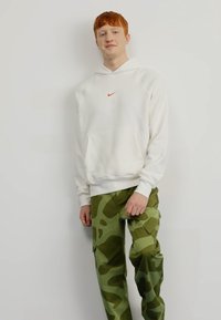 Balta striukė su raudonu "Nike" logotipu, turi kengūros kišenę ir palankiai pritaikytus rankogalius. Žali kamufliažo cargo kelnės su didelėmis šoninėmis kišenėmis ir laisvu siluetu.