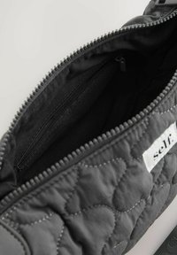 Pikowana czarna torba materiałowa z zamkiem rozchylonym, ukazującym pustą wnętrze oraz kieszeń wewnętrzną na zamek, oznaczoną jako „self”.