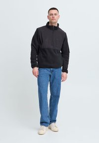Schwarzer Fleece-Pullover mit hohem Kragen und halbem Reißverschluss, ausgestattet mit einer Brusttasche. Getragen mit blauen Jeans im Straight-Leg-Schnitt und cremefarbenen Sneakern.