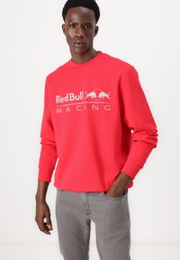 Mężczyzna w jaskrawoczerwonej koszuli z długim rękawem z białym logo „Red Bull Racing”, w jasnoszarych dżinsach, stojący z jedną ręką w kieszeni.