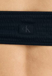 Juodas rhinmanis audinys su lygia tekstūra, su iškilusia juoda "cK" logotipo etikete. Viršutinė kraštinė yra elastinga.