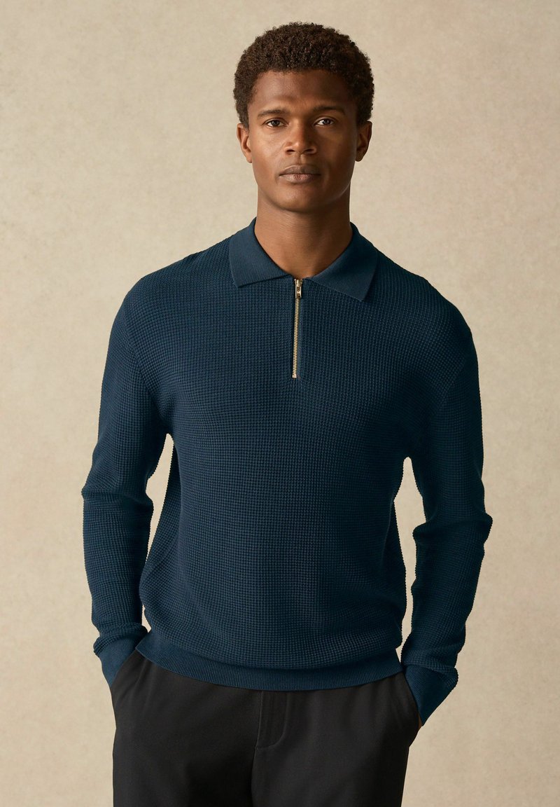 Navy pullover tekstuurilla, jossa on vetoketjukaulus ja pitkähihaiset hihat, istuva siluetti, yhdistettynä mustiin housuihin.