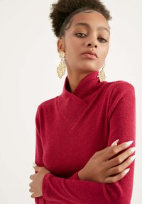 Maglione rosso a coste con colletto alto e ripiegato. Le braccia del modello sono incrociate, evidenziando unghie lunghe. Orecchini in oro texture aggiungono contrasto.