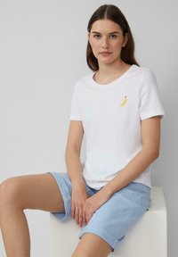 Witte katoenen T-shirt met een gele banaan geborduurd op de linkerkant van de borst, gedragen met lichtblauwe shorts met opgerolde mouwen.