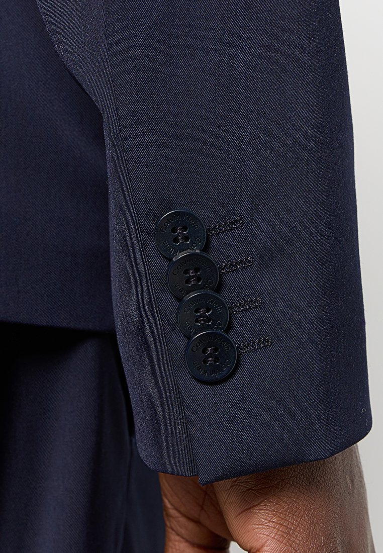 Poignet de blazer marine avec quatre boutons noirs arborant la marque Calvin Klein ; texture lisse avec des détails de couture sur mesure.