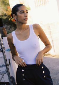 Femme aux cheveux tirés en arrière, portant un débardeur blanc et un pantalon noir à boutons, debout à l'extérieur près d'un fauteuil roulant par une journée ensoleillée.