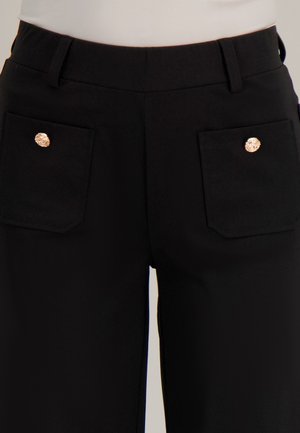 Zwarte broek met twee achterzakken die zijn vastgemaakt met decoratieve gouden knopen, gedragen met een wit topje dat is ingestopt.