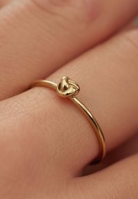 Bague en or avec un anneau mince et lisse, et un petit détail accentué en noeud sur le dessus. La surface est brillante, réfléchissant la lumière de manière subtile.