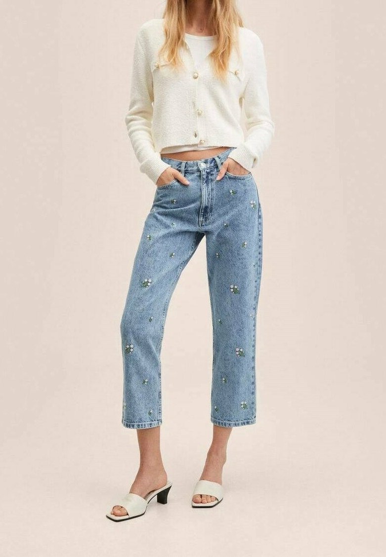 Gilet léger en crème avec boutons, associé à un jean bleu orné de broderies florales. Des sandales à talons ouverts blanches complètent le look.