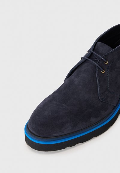 Chaussure en daim bleu marine avec un bout arrondi, présentant une bande bleue contrastante le long de la semelle et des œillets dorés pour les lacets.