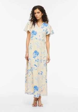 Žena s kudrnatými vlasy, oblečená v béžových maxi šatech s modrým květinovým potiskem a rozevlátými rukávy, stojící před bílým pozadím.