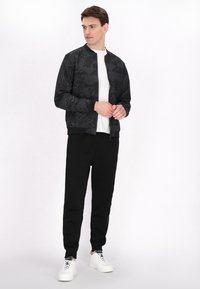 Giacca bomber nera con motivo geometrico, polsini e colletto a coste. Indossata sopra una maglietta bianca, abbinata a pantaloni della tuta neri e sneaker bianche.