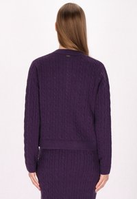 Lila kabelstickad cardigan med långa ärmar, rund halsringning och en texturerad ribbad fåll. Diskret logga vid halsen.