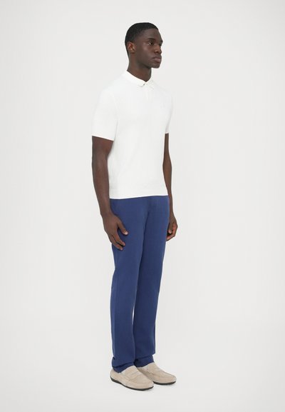 Biela rebrovaná polokošeľa s krátkym rukávom, tmavomodré nohavice a béžové slip-on topánky. Model stojí vzpriamene pred jednofarebným pozadím.