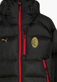 Veste matelassée noire avec fermetures éclair rouges, logo Puma doré sur la poitrine gauche et emblème AC Milan 1899 sur la poitrine droite.