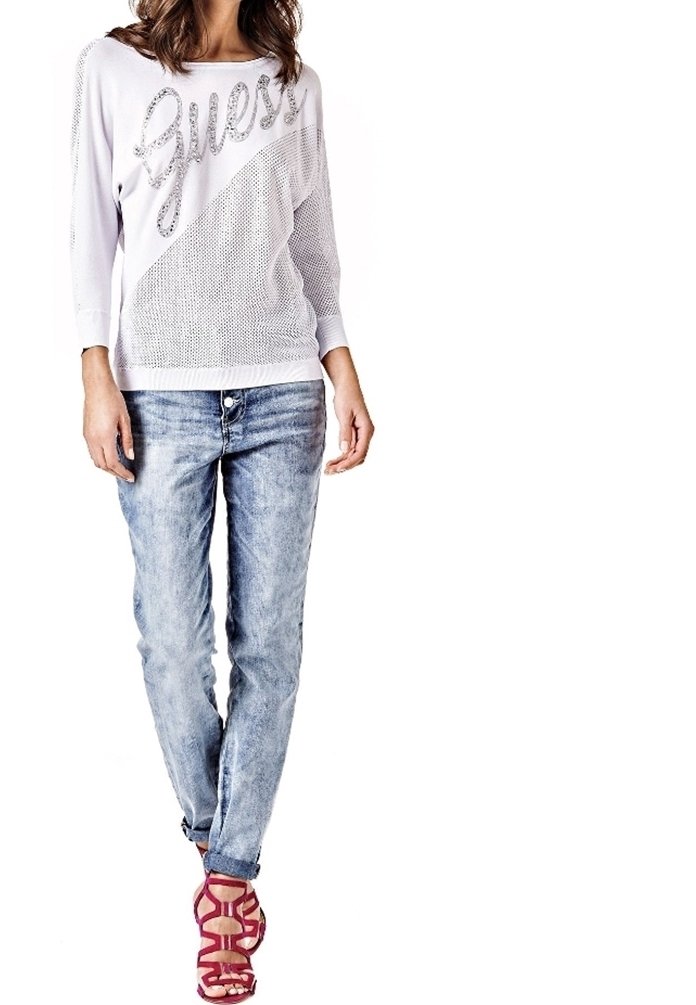 Haut à manches longues blanc avec une texture en maille et "Guess" imprimé en lettres scintillantes, assorti à un jean clair déchiré et des sandales à lanières roses.
