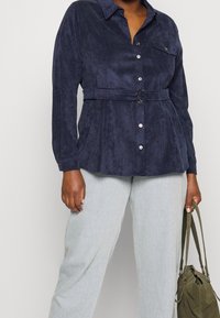 Navyblå button-down skjorta med en mjuk, texturerad yta, bältad vid midjan med en silverring. Har en enda bröstficka och långärmar.