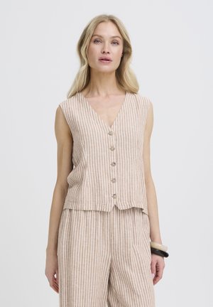 Lyshåret kvinde iført en beige og hvid lodret stribet ærmeløs knap-op top og matchende vidde bukser, stående mod en hvid baggrund.
